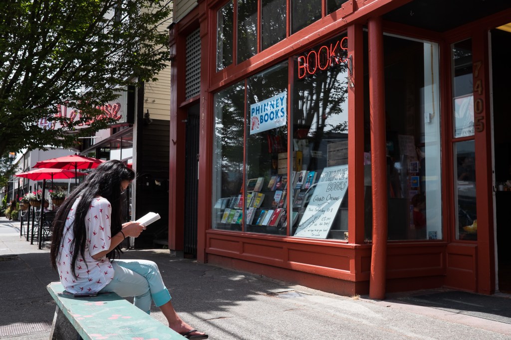 Dark-haired woman reading on a bench in front of a bookstore. Signs on the bookstore read "Phinney Books" and "BOOKS" in neon lights.